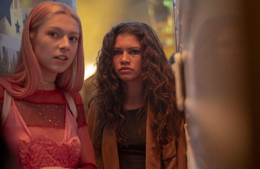 Rue, played by Zendaya Coleman, and Jules, played by Hunter Schafer, hide in-between carts at the town carnvial.  