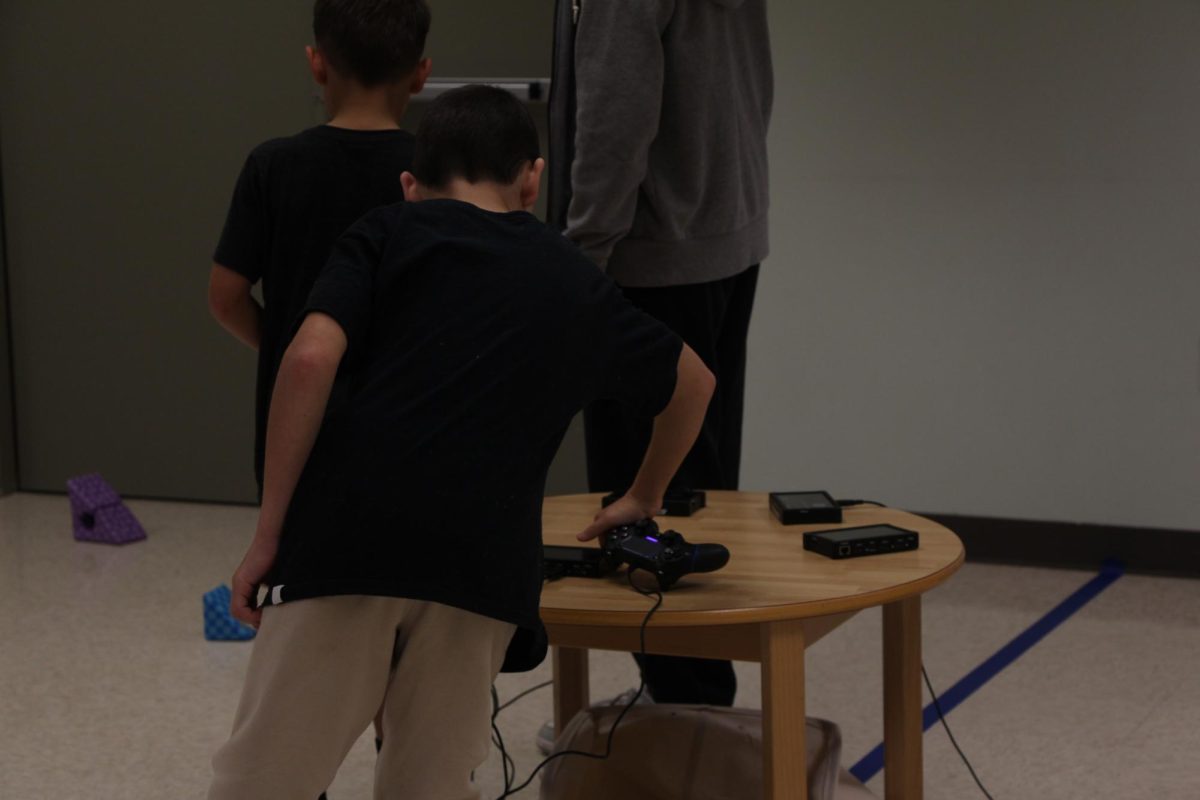 Taking control. A student grabs one of the controllers for the robots off the table. In the center of the room lies the table with all the controllers to drive the robot. Each time a new kid went to drive a robot or put one away, they came back to this table.